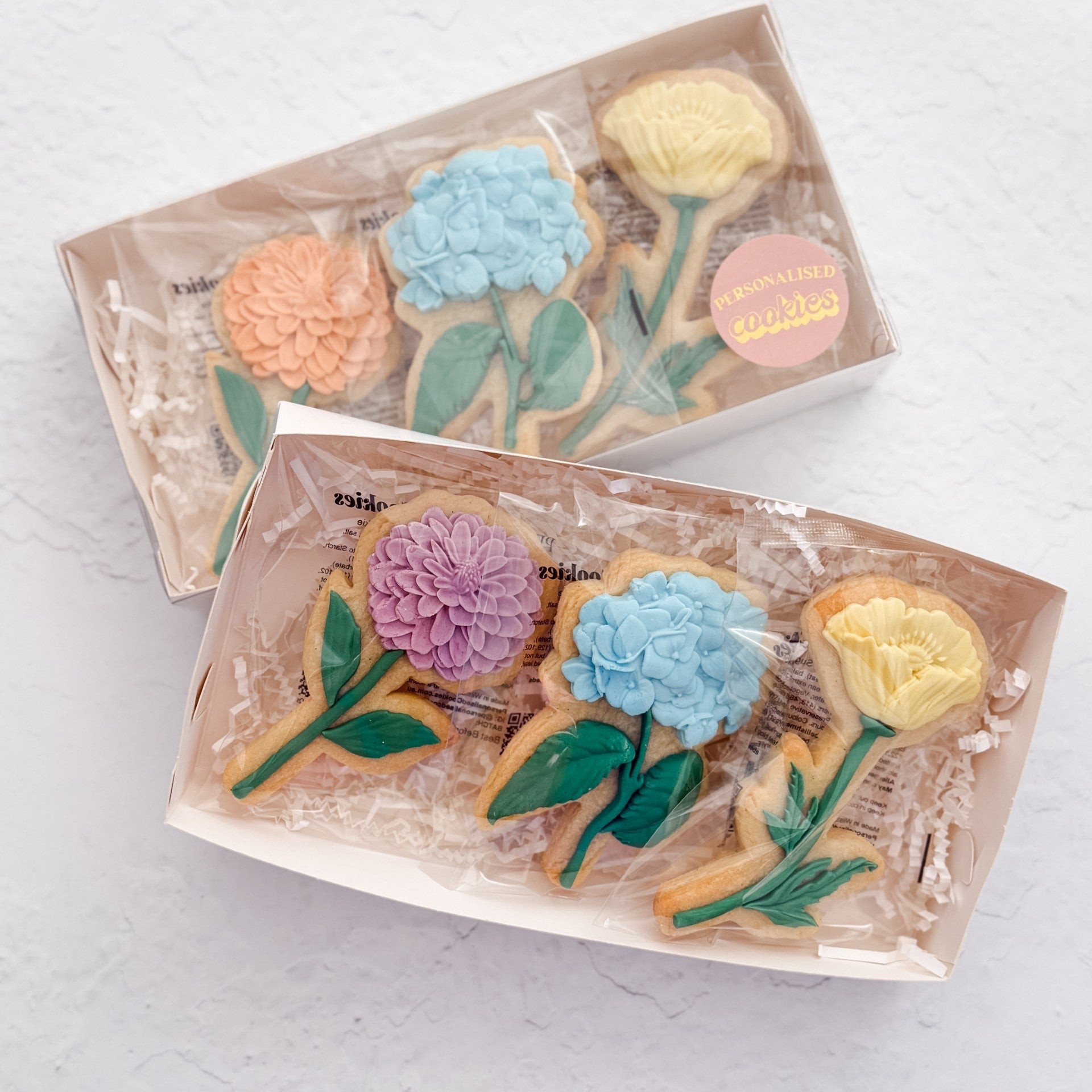 Two clear plastic gift boxes containing decorated flower-shaped cookies, arranged on a light textured white background. The cookies are decorated with pastel icing in pink, blue, yellow, and green, and a small round sticker on the box reads 'Flourish with a Winkles'.