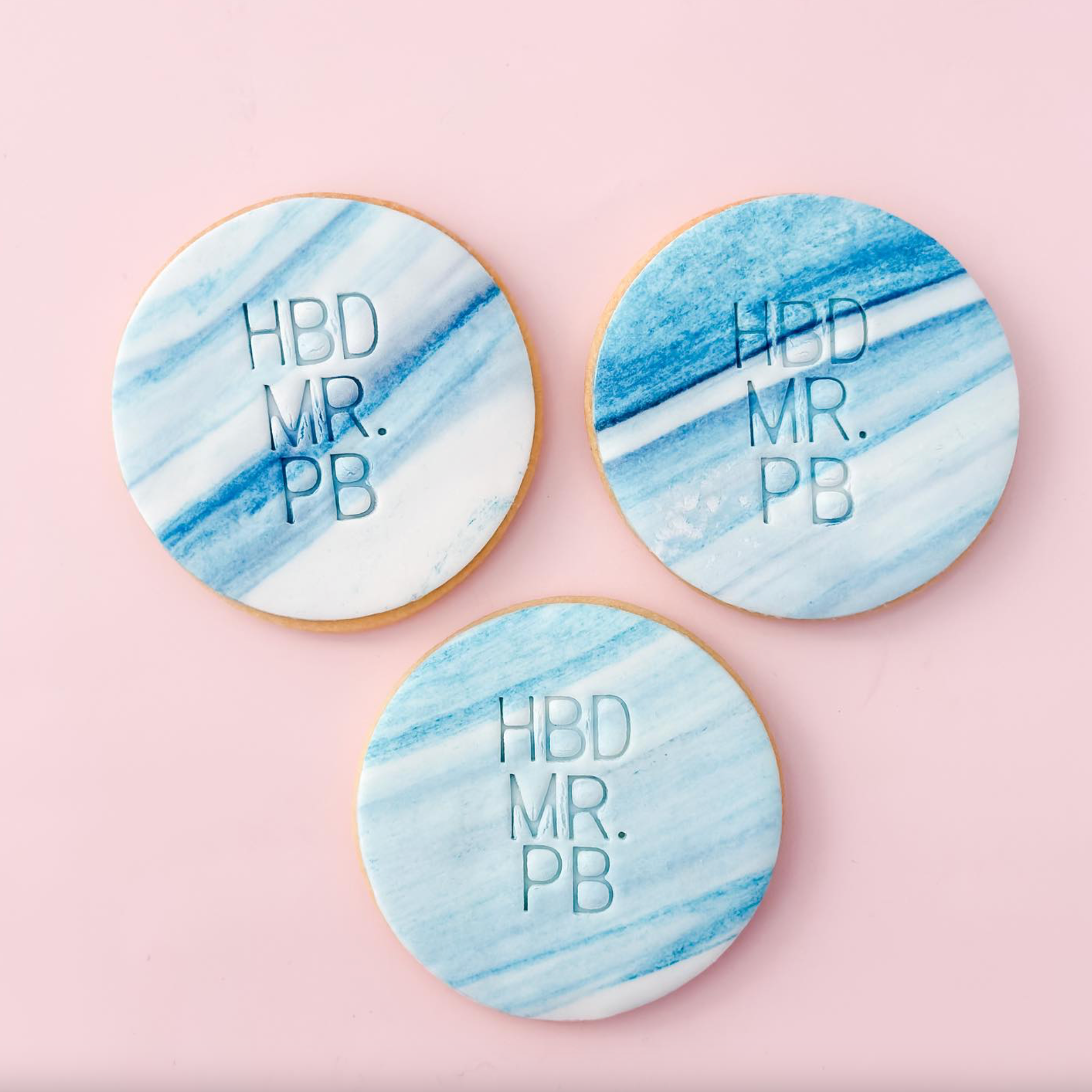Three cookies with blue and white marble design and 'HBD MR. PB' text on a pink background