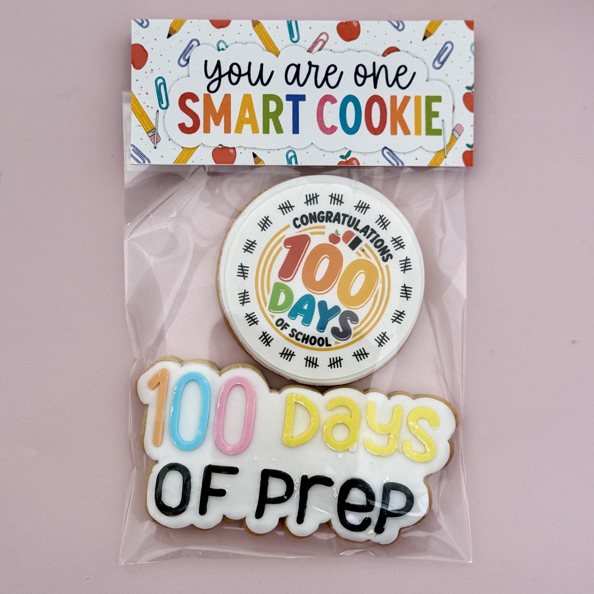 A packaged cookie set on a dusty pink background, featuring a rectangular card that reads 'you are one SMART COOKIE' and two decorated cookies: a round cookie saying 'Congratulations on 100 Days of School' and a cloud-shaped cookie reading '100 Days OF Prep'.