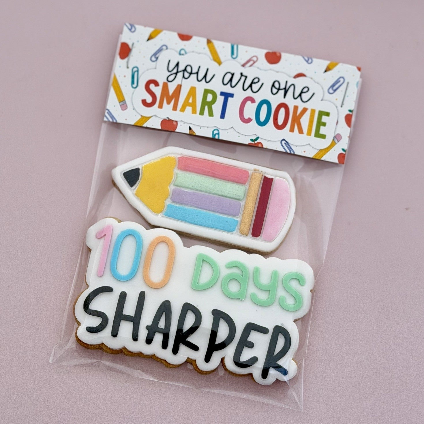 A packaged sugar cookie with a printed header card that reads 'you are one SMART COOKIE.' The cookie is shaped like a pencil and a stack of colorful number tiles spelling '100 days SHARPER,' shown on a soft pink background.