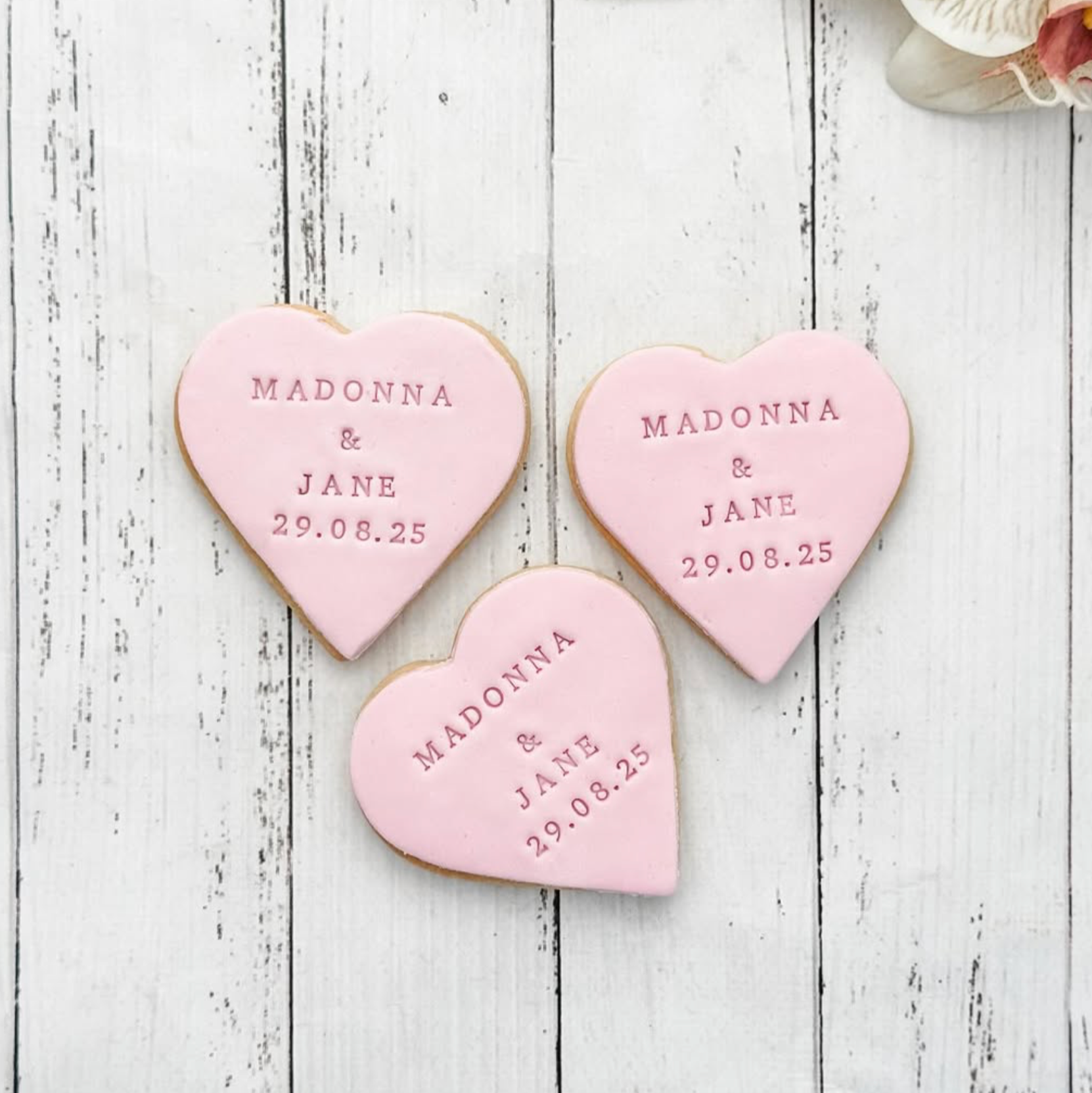 Heart-shaped cookies with personalized names and date on a wooden surface with flowers.