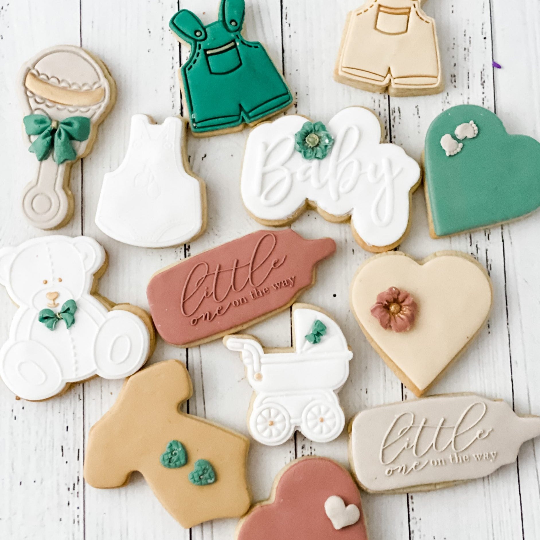 A variety of decorated cookies designed to look like baby items such as a stroller, onesies, hearts, and baby rattle, primarily in neutral colors.
