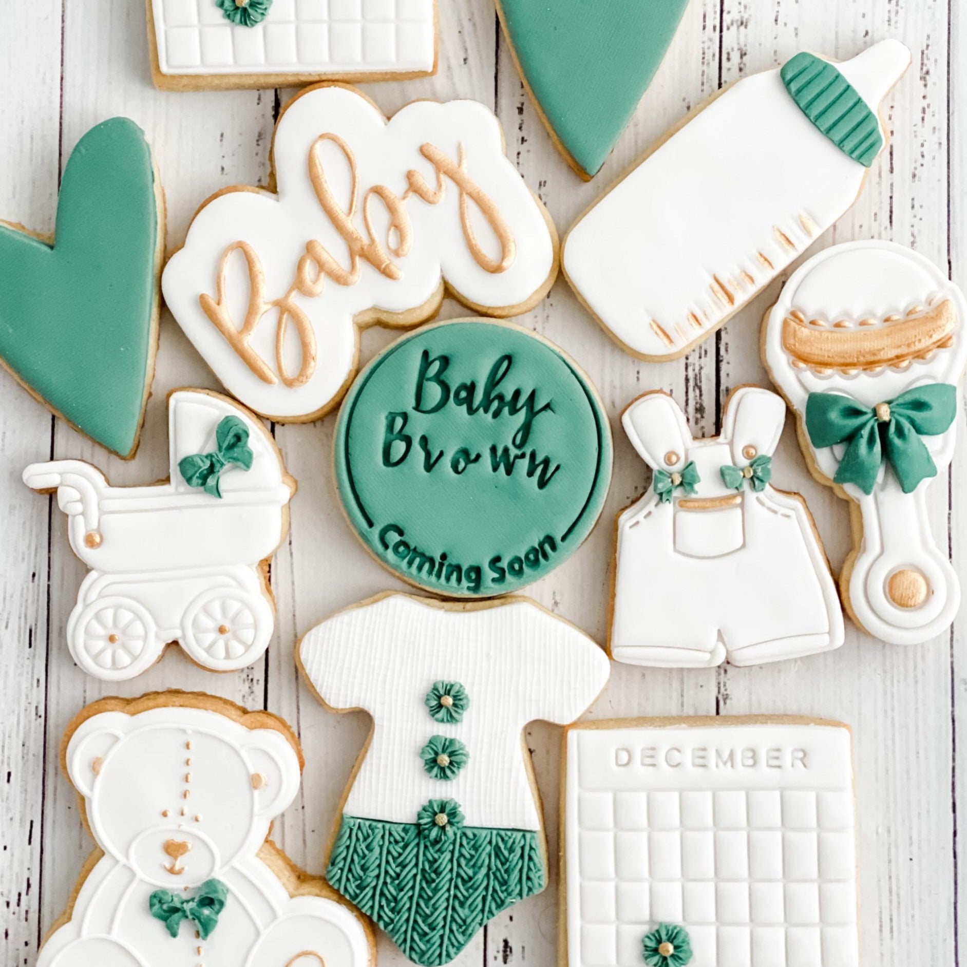 A dozen of variously shaped cookies decorated with baby-themed designs such as a baby shower cake, calendar with due date, onesie, bear, pram, hearts, and text.