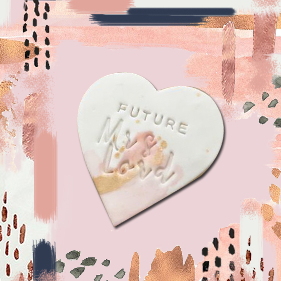 A heart-shaped cookie with the words 'FUTURE Mrs. [last name]' written on it, presented on a pastel-colored background with abstract designs.