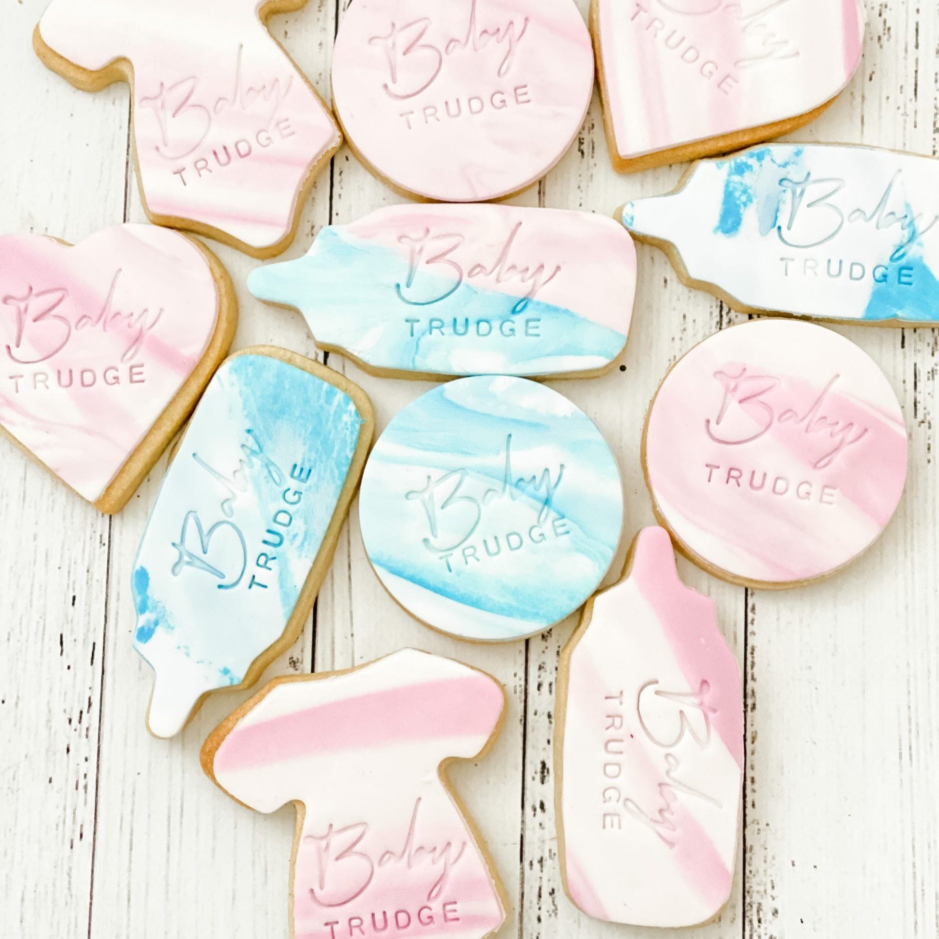 A variety of cookies with baby-themed shapes such as onesies and bottles, decorated in pastel colors with the words 'Baby Trudge' stamped on them.