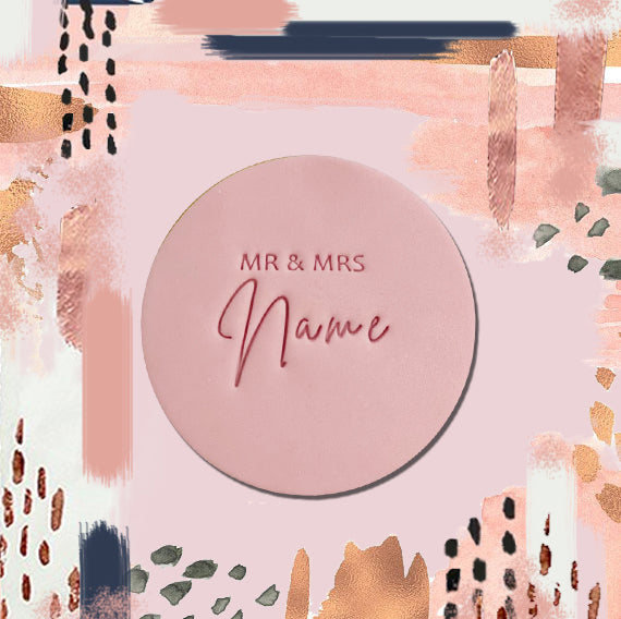 A custom sugar cookie with 'MR & MRS' and a placeholder for a name, presented on a round pink plate with a decorative background.