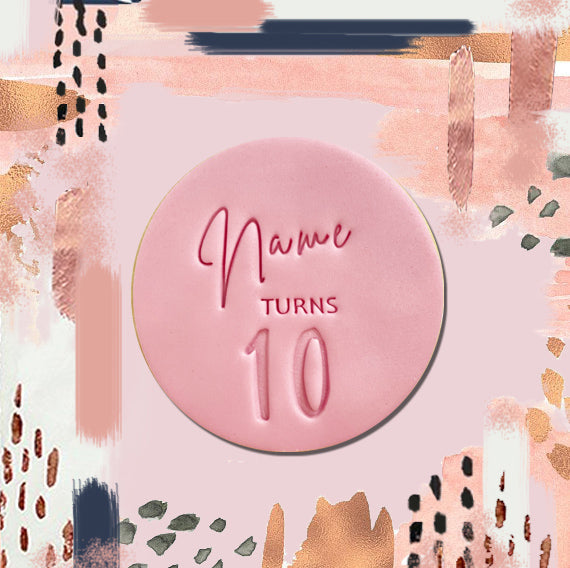 A round cookie with pink fondant icing, featuring custom text 'Name turns 10' printed on it, against a patterned background.