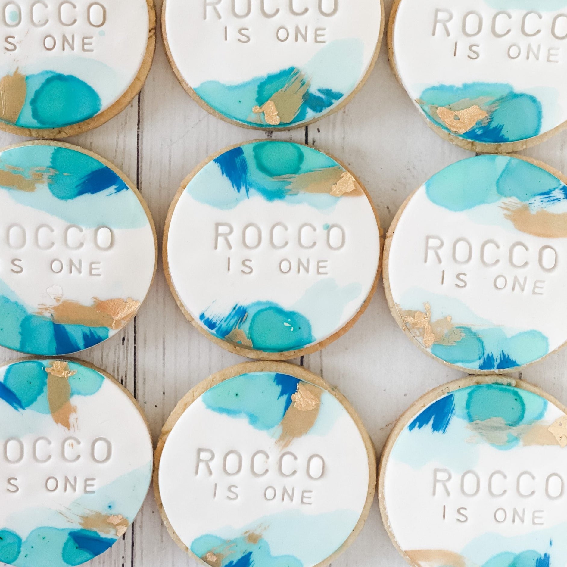watercolour cookies in shades of blue, teal and gold with Rocco embossed into fondant. placed on a white background 