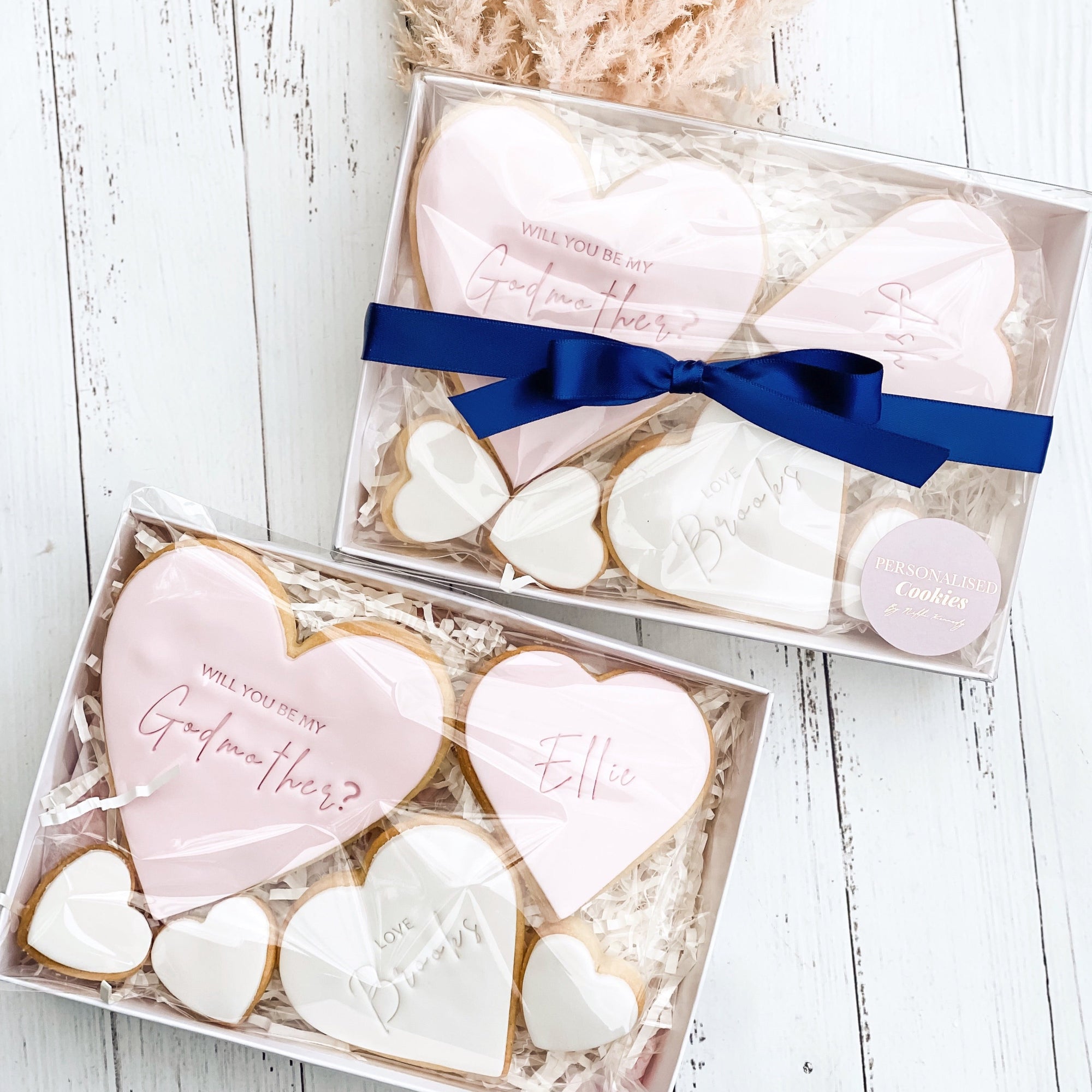 A box of personalized heart-shaped cookies, with one cookie personalized with a name, and three mini hearts. The box is adorned with tissue paper, has a window facing the cookies, and is tied with a navy ribbon.