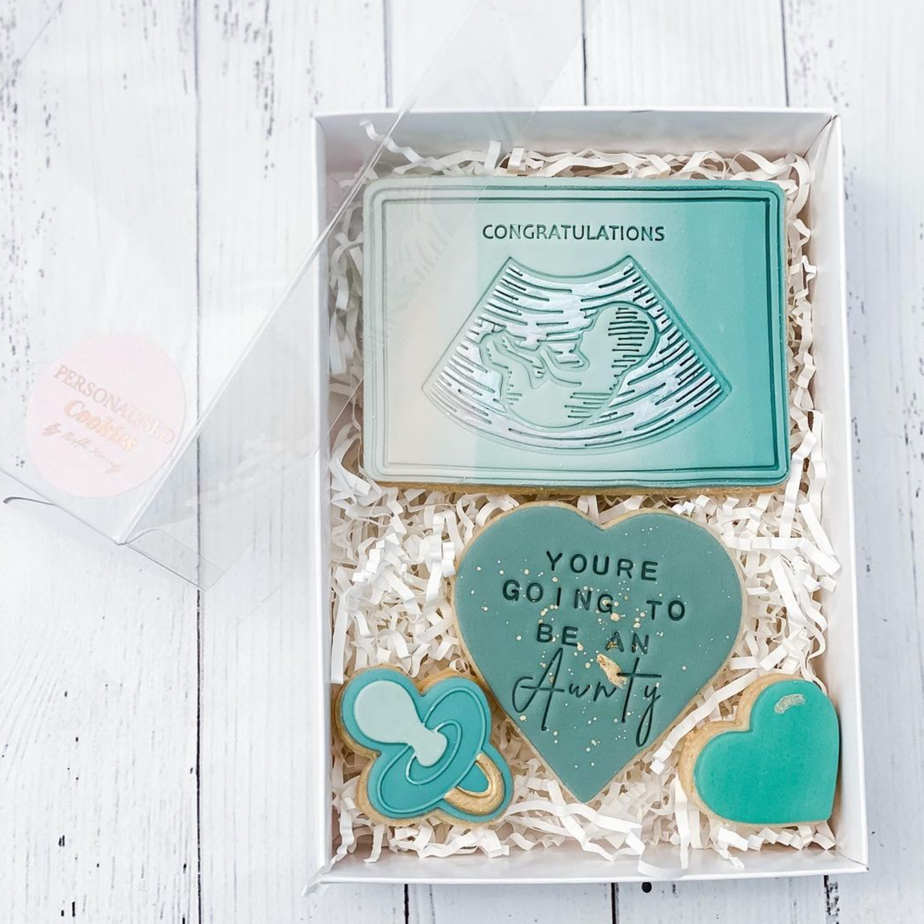 A gift box containing a large cookie with a heart shape, two smaller cookies, and a card with an ultrasound scan image. The cookies are teal-colored with white icing for the heart and text, presented on a white shredded filling in a clear box, with a pink ribbon on top.